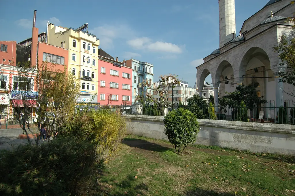 Firuz Ağa Camii'nin caddeye bakan yanı; önde çimenlik ve çalılar, alçak avlu duvarı, arkada renkli cepheli binalar.