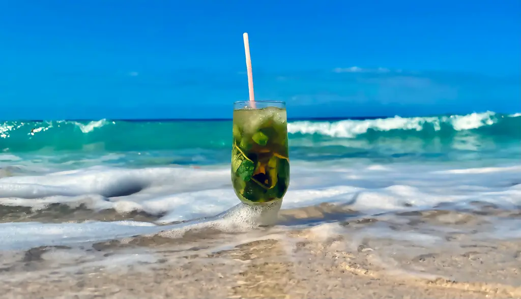 Kumsalda duran uzun bardakta mojito; içinde nane yaprakları ve buz, arkasında köpüren dalgalar ve turkuaz deniz.