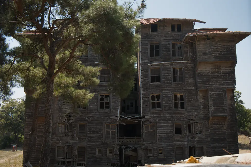 Büyükada Rum Yetimhanesi 2026 yazında; koyu ahşap cephe, kırık pencereler ve önde çam dalları.