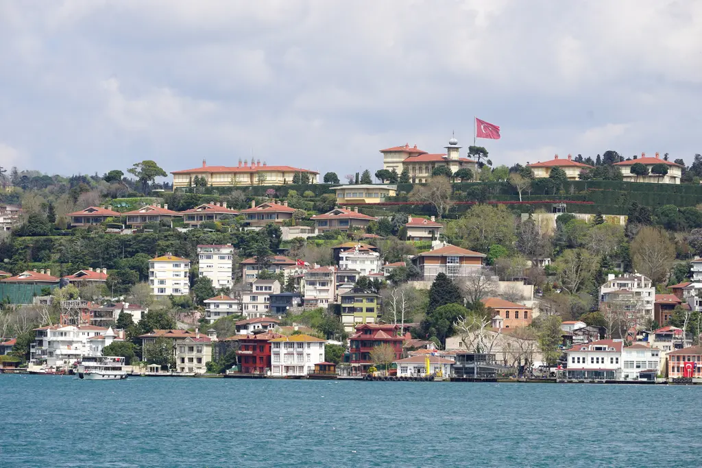 Çengelköy'e Boğaz'dan bakış; kıyıda yalılar, yamaç boyunca sıralanan kiremit çatılı evler ve tepede bayraklı köşk.