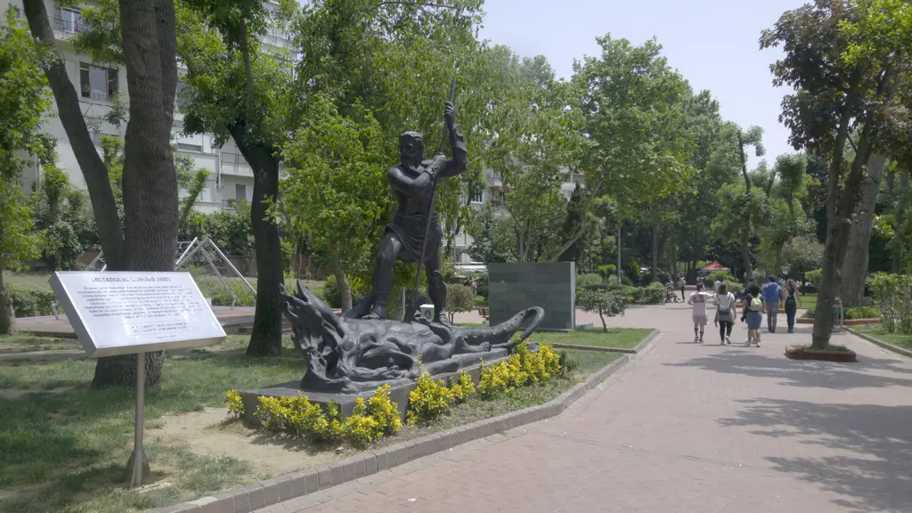 Maçka Demokrasi Parkı'nda bronz bir heykel; kılıç kaldıran figür, ayağının dibinde ejderha benzeri yaratık, sarı çiçekli tarh ve bilgi levhası.