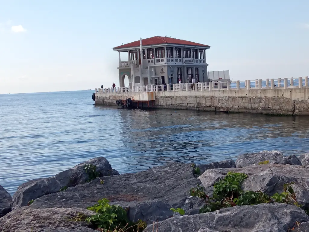 Moda İskelesi; ön planda dalgakıran kayalar, taş iskele yolu, kiremit çatılı bina ve mavi deniz.