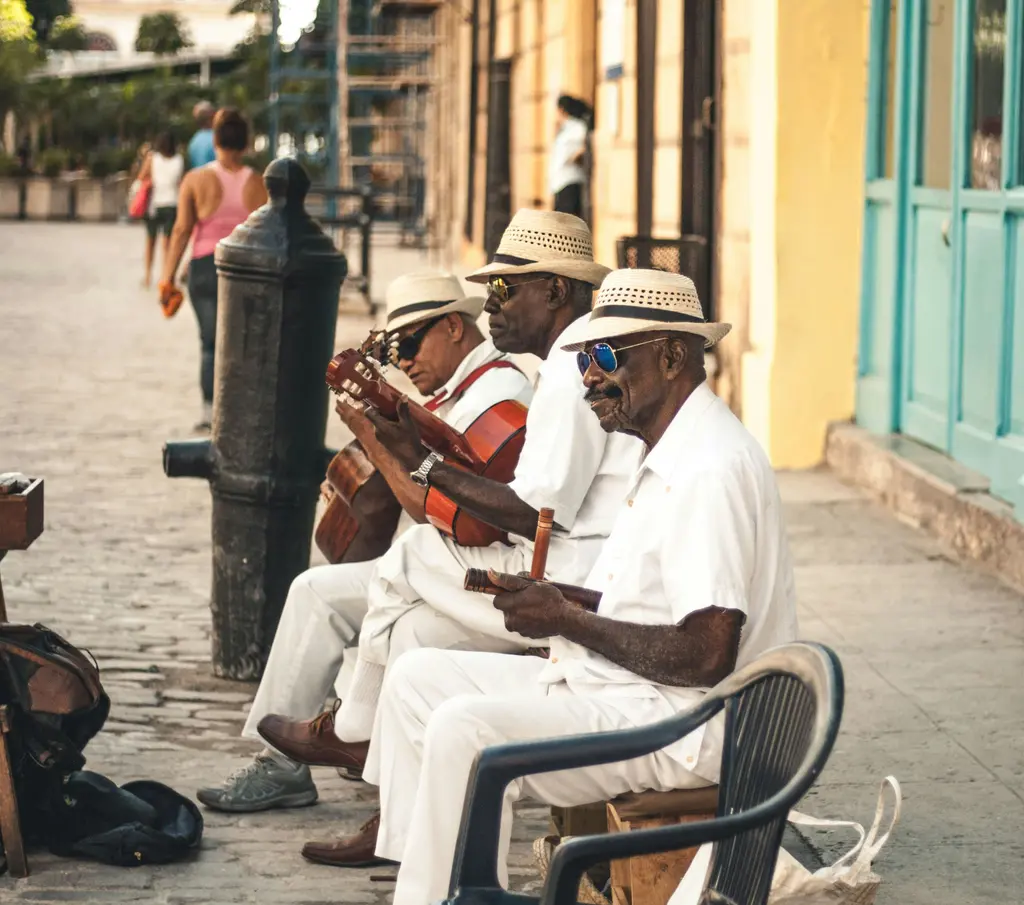 Havana'da bir kaldırımda oturan üç müzisyen; beyaz gömlek ve hasır şapkalarıyla gitar ve perküsyon çalarken arkalarında sarı kolonyal bir bina.
