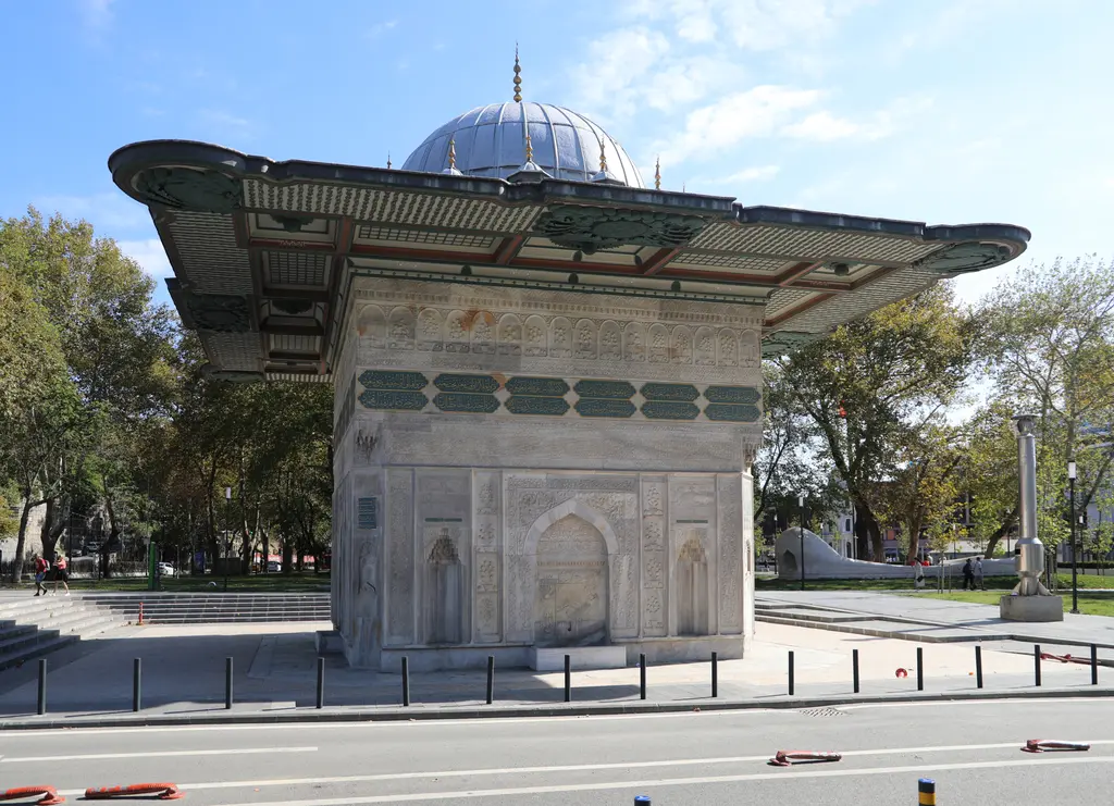 Tophane Çeşmesi'ne köşeden bakış; iki cephe, köşede kabartmalı yüzey, üstte yeşil zeminli yazı kuşağı ve geniş saçak.