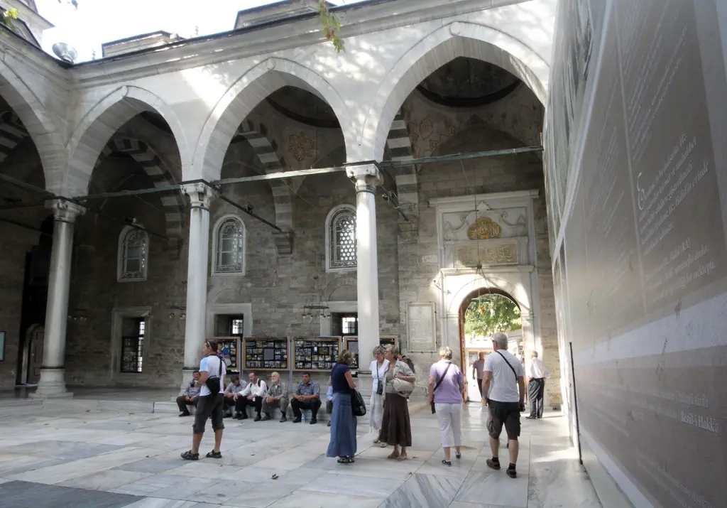 Eyüp Sultan Camii'nin iç avlusu; kubbeli revaklar, mermer sütunlar, kemerli kapılar ve dolaşan ziyaretçiler.