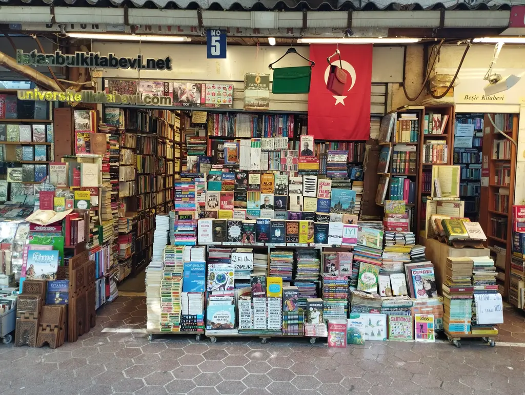 Sahaflar Çarşısı'nda tavana kadar kitapla dolu bir dükkân ağzı; ortada asılı büyük Türk bayrağı, önde tekerlekli standlarda "2. el ucuz kitaplar" yığınları ve yeşil sarı tabela.