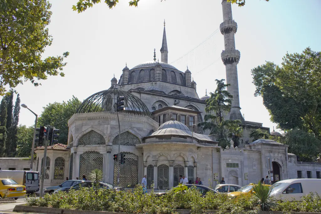 Üsküdar'da Yeni Valide Camii; kubbe, iki minare, önde kafes kubbeli türbe ve sebil, caddedeki araçlar.