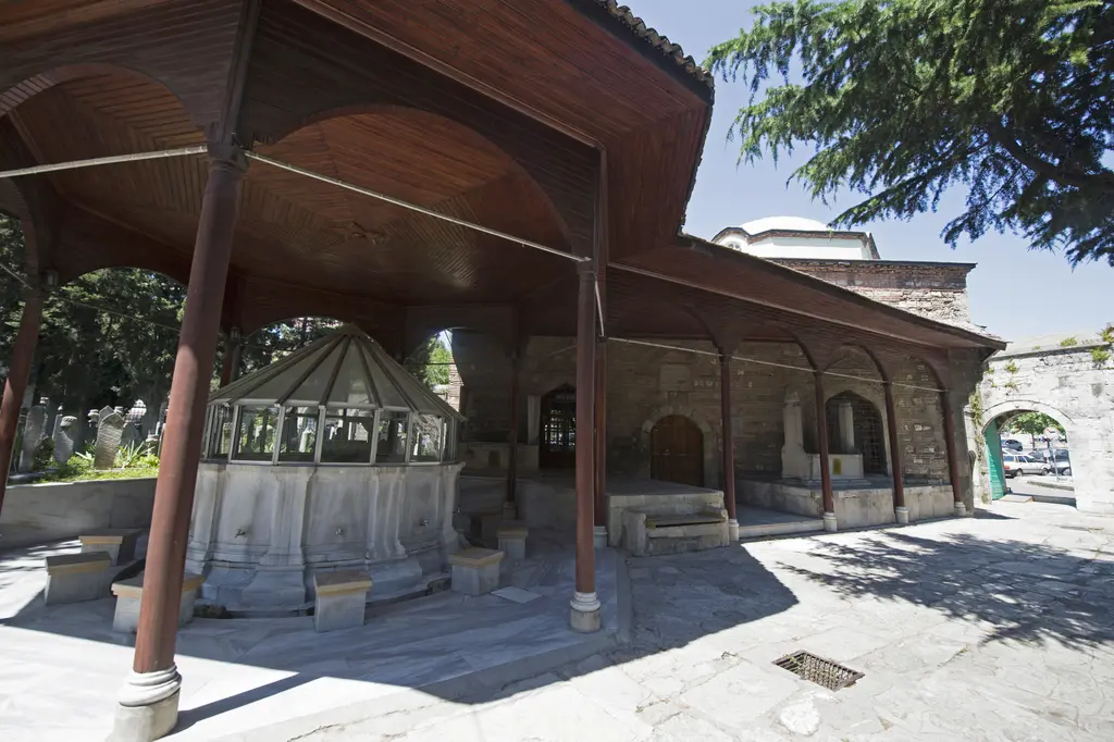 Şadırvan; ahşap direklere oturan geniş saçak altında mermer hazne, çevresinde musluklar ve arkada hazire ile avlu.