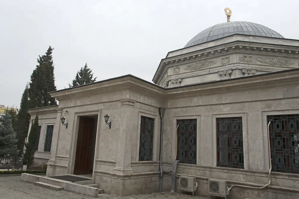 II. Mahmud Türbesi'nin kuzey yüzü; türbeye bitişik tek katlı mermer bölüm, ahşap kapı, şebekeli pencereler, arkada kubbe ve yanda serviler.
