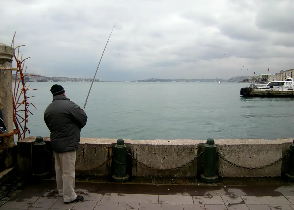 Ortaköy kıyısında olta atan bir balıkçı; taş korkuluk, gri Boğaz suyu, uzakta vapur ve karşı kıyı.