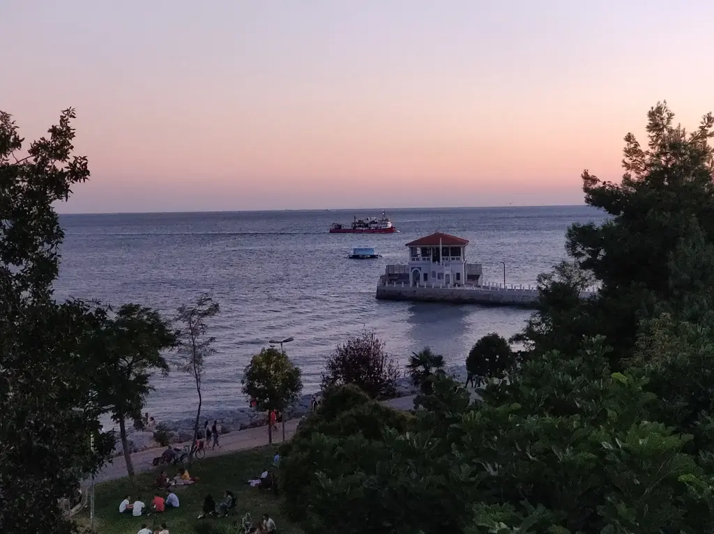 Moda Parkı'ndan iskele; ağaçların arasından deniz, uzakta iskele binası, çimende oturan insanlar.