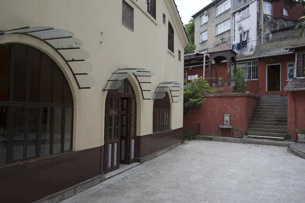 Balat'ta Panayia Balino Kilisesi avlusu; krem boyalı kilise cephesi, kemerli büyük pencereler, taş döşeli avlu ve arkada merdivenli kırmızı yapılar.
