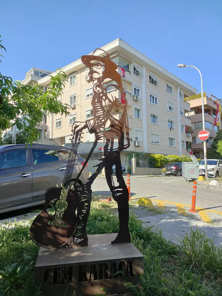Moda'da Cem Karaca heykeli; gitarlı müzisyen silueti, metal levhadan kesilmiş gövde, çimen ve arkada apartman.