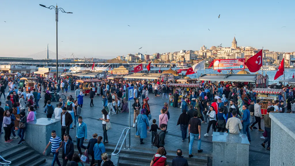 Eminönü kıyısında kalabalık; rıhtım boyunca yürüyen insanlar, bayraklar, tezgâhlar ve karşıda Galata Kulesi.