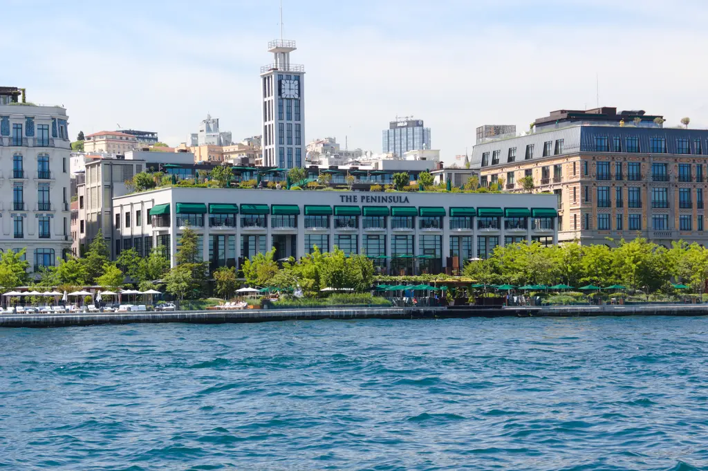 Denizden The Peninsula Istanbul; yeşil tenteli eski yolcu salonu binası, önünde ağaçlar ve kıyı, arkada eski hanlar ve kule.