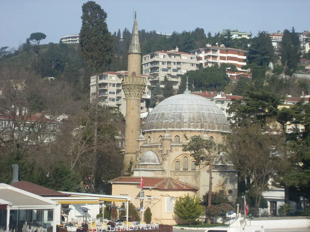 Bebek Camii'nin kubbesi ve minaresi; gri kubbe, kemerli pencereler, arkada yamaçtaki apartmanlar ve ağaçlar.
