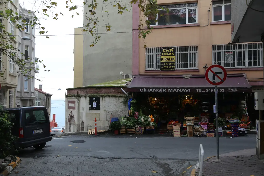 Cihangir'de köşedeki manav; tezgâhlarda meyve ve sebzeler, yokuş yukarı sokak ve yanında sarı cepheli apartman.