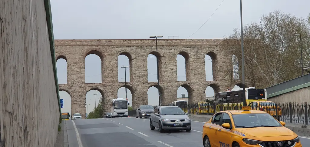 Bozdoğan Kemeri'nin uzun cephesi; altta büyük yuvarlak kemerler, üstte dar kemer sırası, sağda gözün altından geçen sarı taksi, önde beton istinat duvarı ve yol.