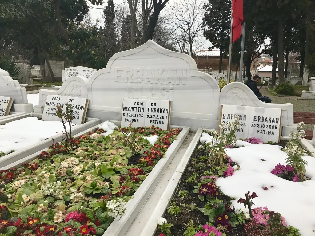 Aile kabristanı; beyaz mermer sandukalar, üzerlerine ekilmiş kırmızı çiçekler, arkada mezar taşları ve ağaçlar.