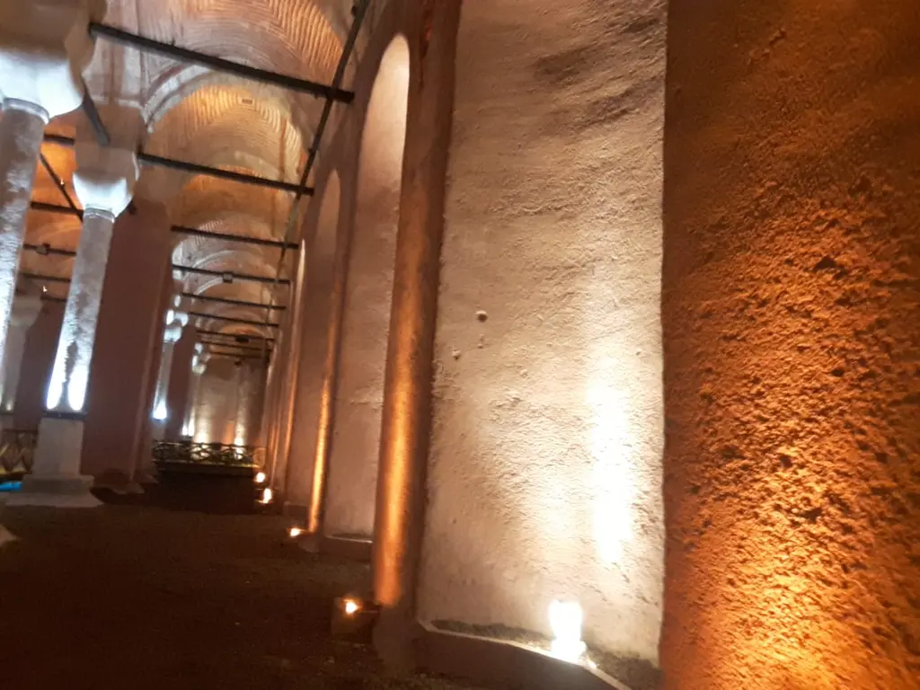 Zeyrek Sarnıcı'nın iç duvarı; turuncu ışıkla aydınlanan kaba sıvalı duvar, kemerli nişler, sütunlar ve zeminde küçük spotlar.