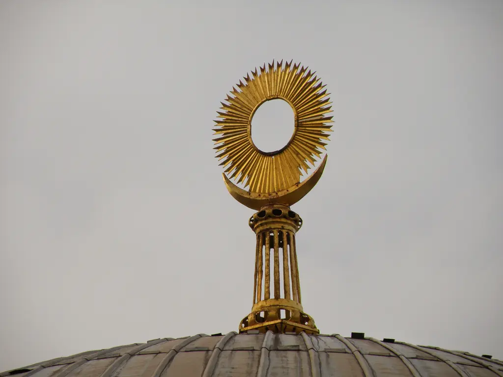 II. Mahmud Türbesi'nin alemi; kubbe tepesinde altın yaldızlı, ışınsal halka biçimli alem, sütunçelik gövde ve arkada bulutlu gök.