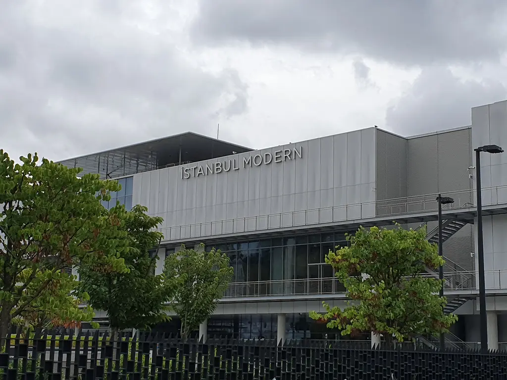 Ağaçların arasından İstanbul Modern'in cephesi; açık gri kaplamada müzenin adı ve bulutlu gökyüzü.