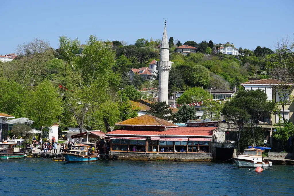 Kanlıca'ya denizden bakış; kıyıda kırmızı tenteli lokanta, tekneler, ağaçlar arasında yükselen ince minare ve sırttaki evler.