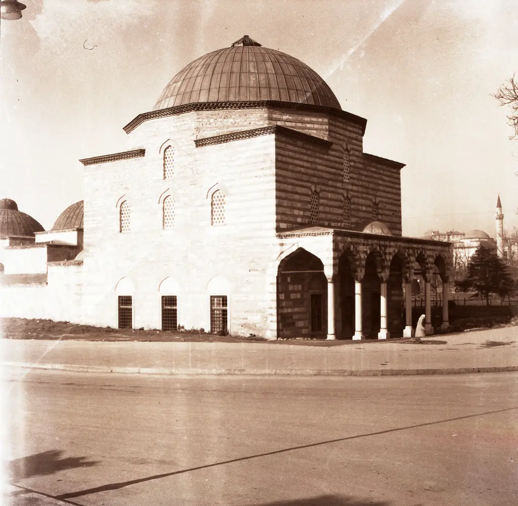 Eski bir fotoğrafta hamam; sıvasız taş ve tuğla cephe, kubbeli sütunlu revak, boş toprak yol ve arkada bir minarenin ucu.