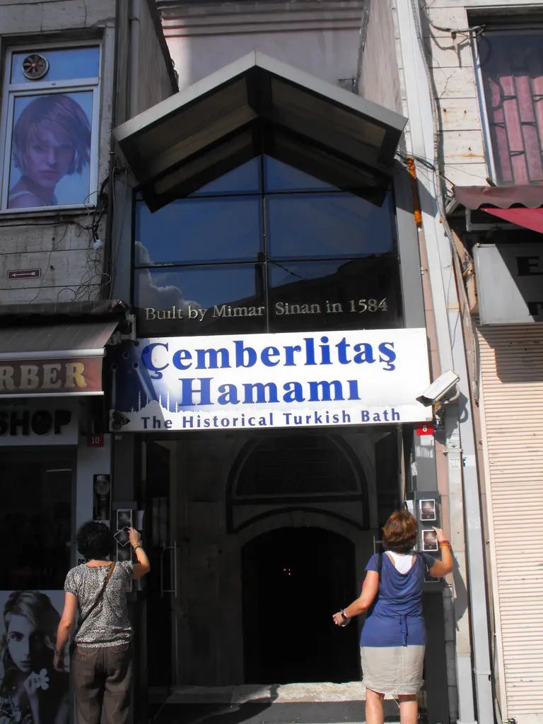 Çemberlitaş Hamamı'nın giriş kapısı; üstünde hamamın adını taşıyan mavi beyaz tabela, kapı üstünde İngilizce bir yazı ve kapıdan giren iki ziyaretçi.