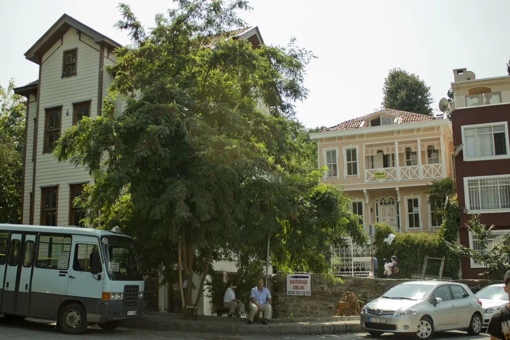 Çengelköy'de yan yana iki eski ev; beyaz ahşap köşk, ağaçlar arasında sarı cepheli ev ve önde minibüs.