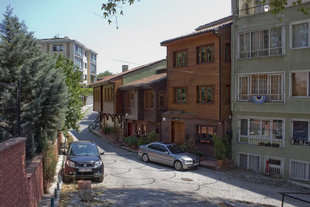 Çengelköy'de yokuş yukarı kıvrılan parke taşlı sokak; ahşap kaplı evler, park etmiş arabalar ve çam ağacı.