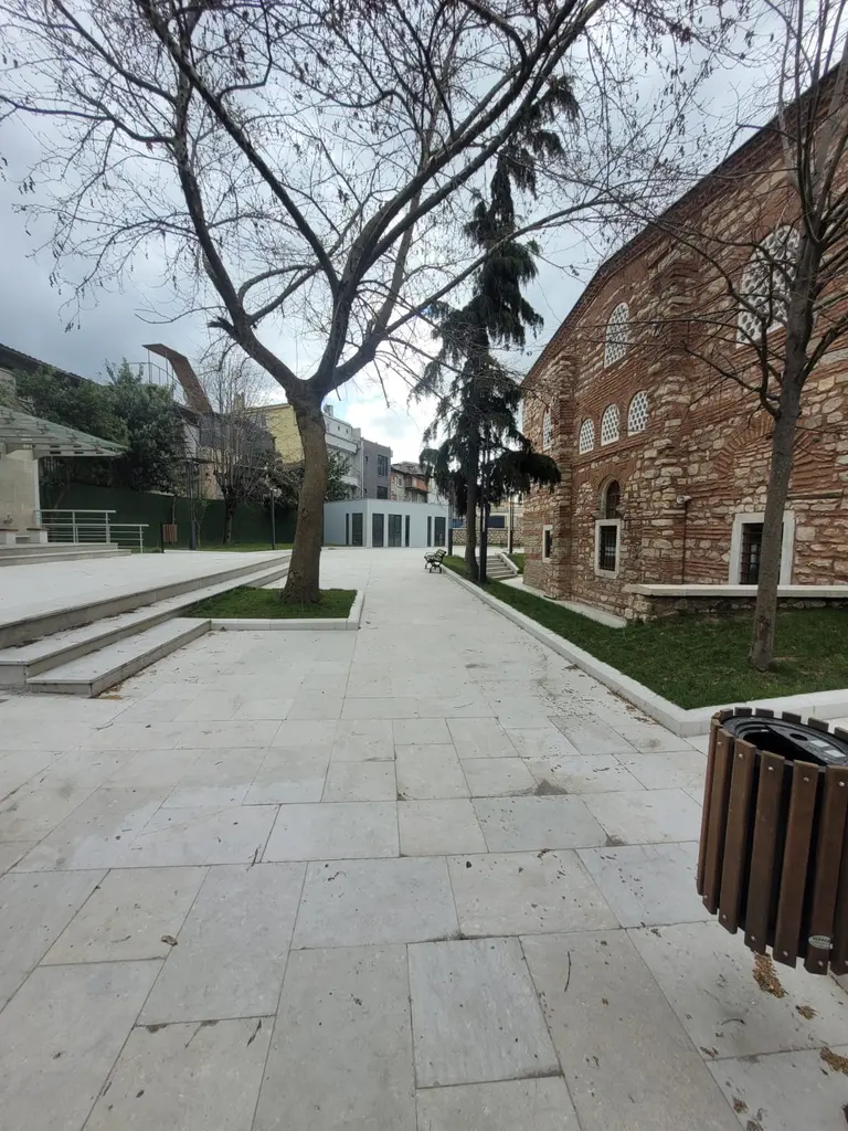 Atik Mustafa Paşa Camii'nin yanından uzanan taş döşeli yol; beyaz taş plakalar, basamaklı set, çimenlik ve genç ağaçlar, sağda tuğla duvarlı cami gövdesi.