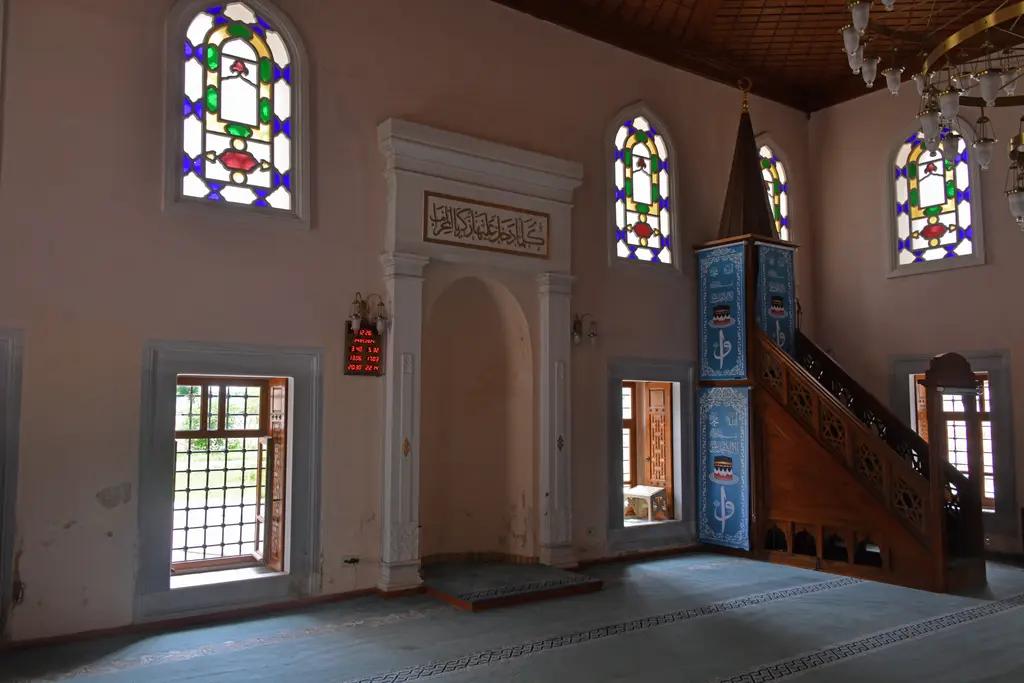 Şah Sultan Camii'nde mihrap ve minber; beyaz mihrap nişi, levha, ahşap minber, mavi örtü ve renkli camlı pencereler.