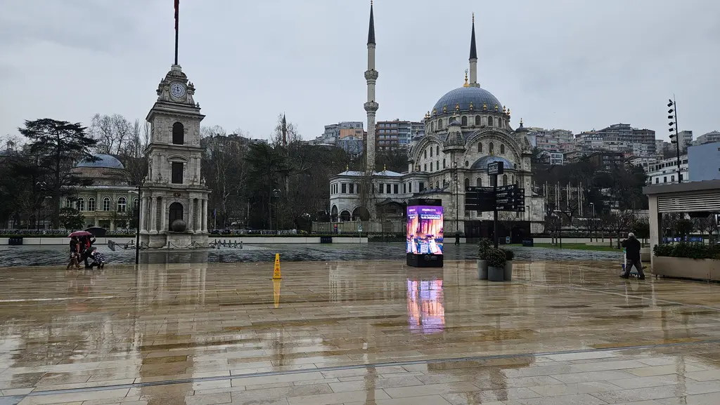 Yağmurlu bir günde Tophane meydanı; ıslak taş zeminde yansımalar, solda saat kulesi, arkada Nusretiye Camii'nin kubbesi ve minareleri.