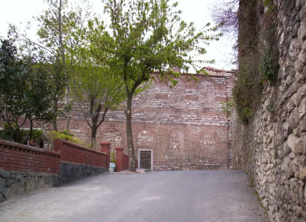 İmrahor İlyas Bey Camii'nin avlu duvarı; yüksek tuğla ve taş örgülü kütle, dipte küçük demir kapı, solda kırmızı tuğla bahçe duvarı ile ağaçlar, önde asfalt.