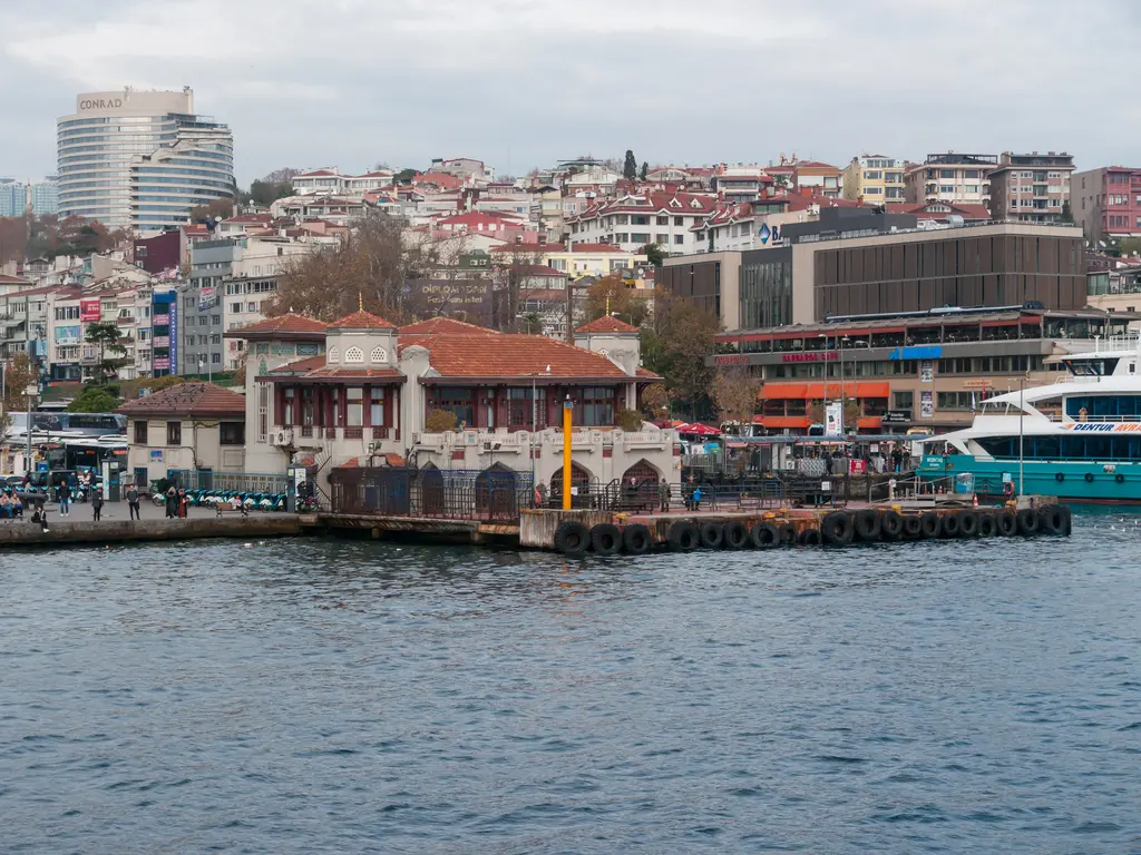 Denizden Beşiktaş; önde iskele, arkada yamaca dizilen binalar, sağda bağlı teknelerle iskele binası ve bulutlu gökyüzü.