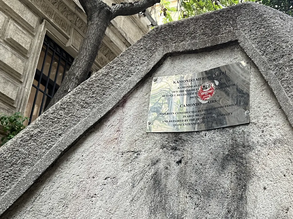 Kamondo Merdivenleri'nin taş korkuluğundaki tanıtım plaketi; Türkçe ve İngilizce yazılar, arkada ağaç gövdesi ve bina cephesi.