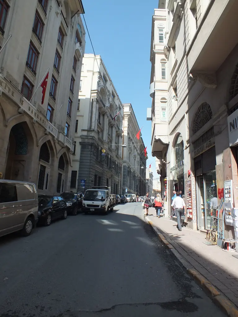Güneşli bir günde Bankalar Caddesi; dar yol, iki yanda yüksek eski binalar, park etmiş arabalar ve uzakta yayalar.