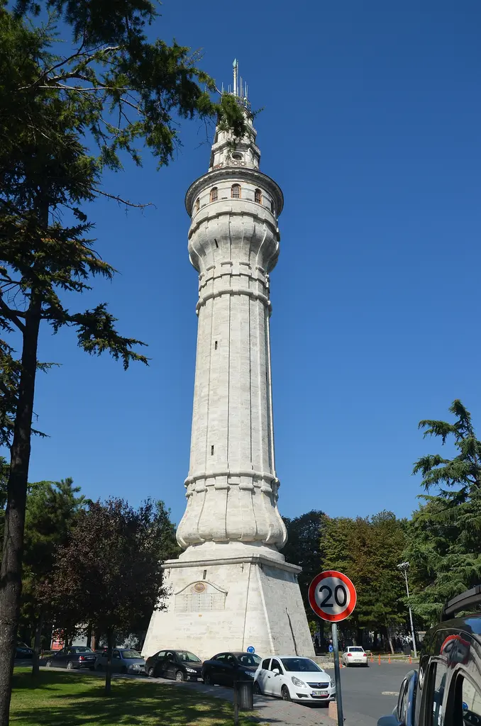 Kampüs yolundan Beyazıt Kulesi; yol kenarındaki ağaçlar, trafik levhası, park etmiş araçlar ve arkada yükselen taş kule.