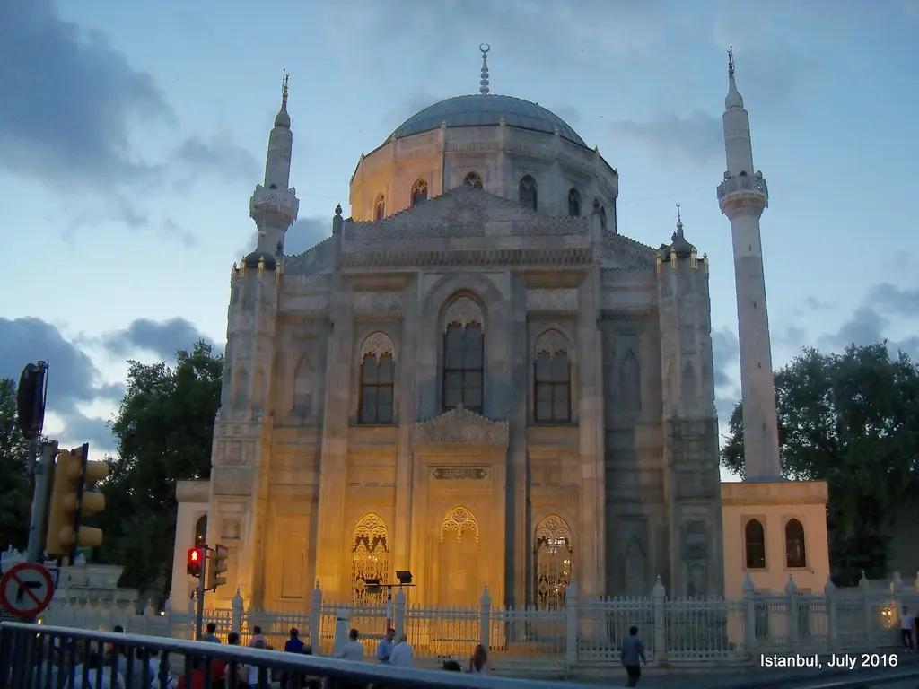 Akşam aydınlatmasıyla Pertevniyal Valide Sultan Camii; ışıklı gotik pencereler, taş cephe, iki minare ve önde trafik lambası.