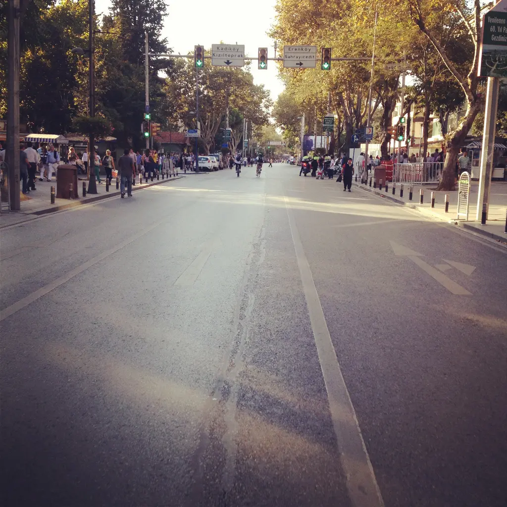 Akşamüstü Bağdat Caddesi; boş asfalt, iki yanda ağaçlar, kaldırımda yürüyenler ve mor tonlu gökyüzü.