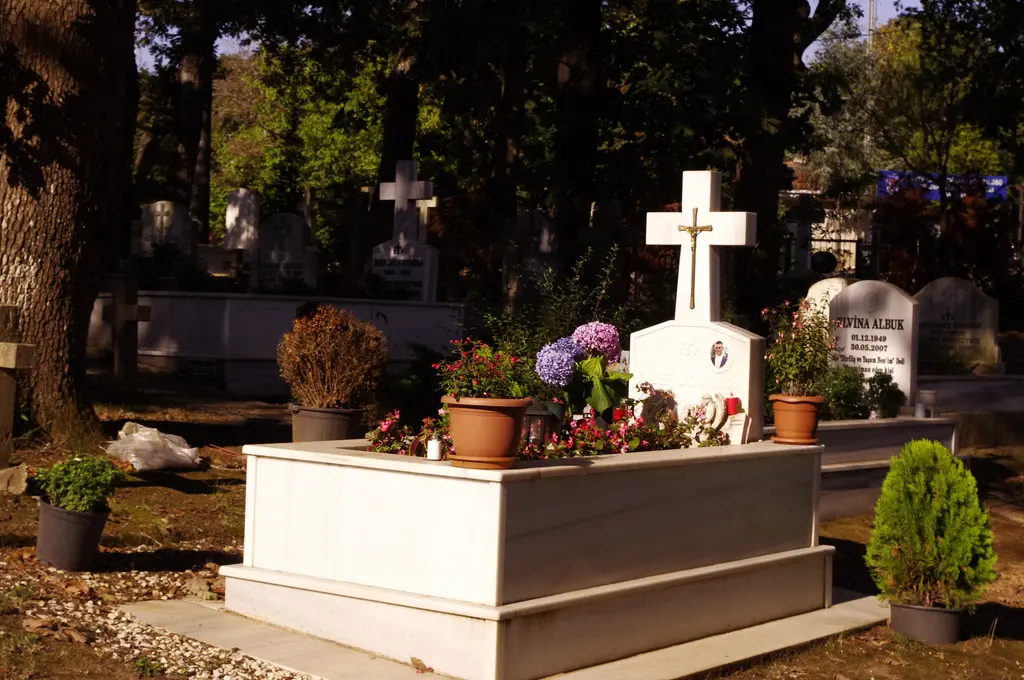 Polonezköy mezarlığında beyaz mermer bir mezar; üstünde haç, saksılarda ortanca ve çiçekler, arkada başka mezarlar ve ağaçlar.