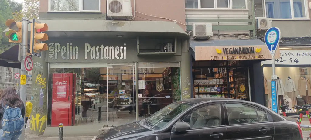 Moda'da Pelin Pastanesi; köşe dükkânın kırmızı tabelası, vitrinler, yanında dükkânlar ve önde park etmiş arabalar.