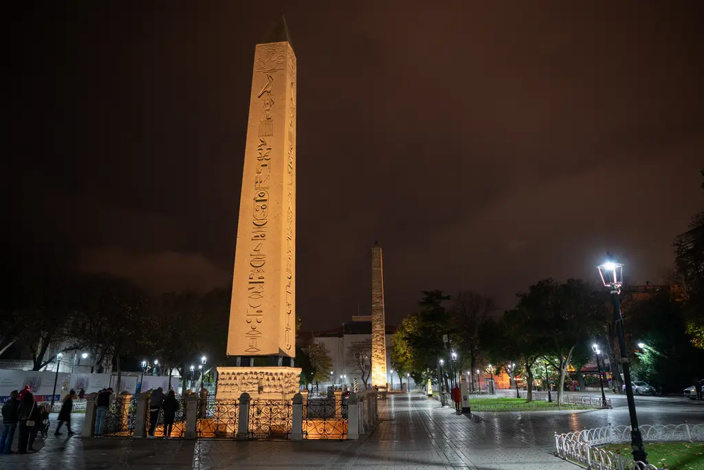 Gece Sultanahmet Meydanı; önde aydınlatılmış Dikilitaş, arkada Örme Sütun, ıslak taş yol, sokak lambaları ve parmaklıklar.