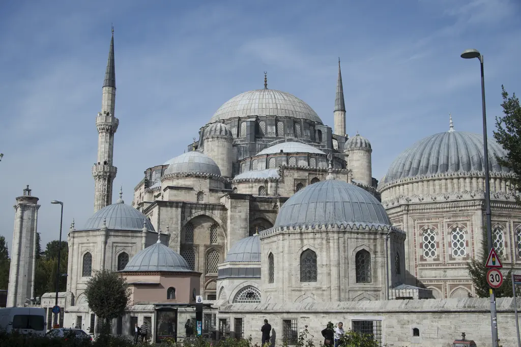 Fatih'te Şehzade Camii sokaktan; iki ince minare, merkezî kubbenin çevresinde basamak basamak alçalan yarım kubbeler ile küçük kubbeler, sağda dilimli kubbeli türbe.