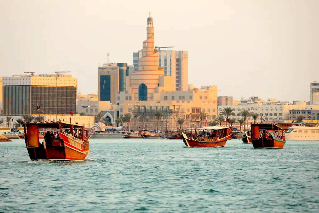 Doha'da helezon biçimli minare; kum rengi sarmal gövdesi, arkasındaki gökdelenler ve önde suda demirlemiş kırmızı-ahşap dhow tekneleri.