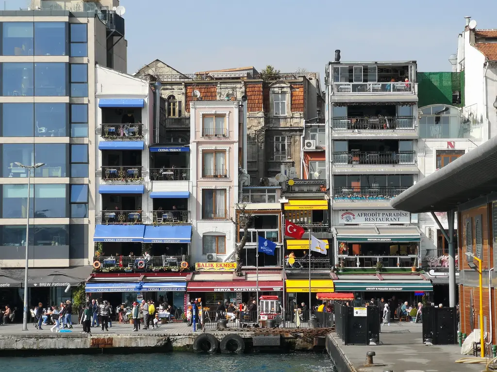 Karaköy rıhtımı; renkli tenteli lokantalar, balkonlu binalar, Balık Ekmek tabelası ve kıyıda yürüyen insanlar.