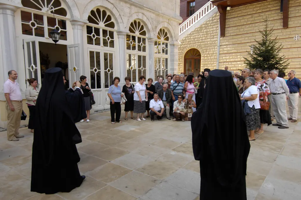 Patrikhane avlusunda siyah cüppeli ve kalpaklı ruhaniler ile çevrelerinde toplanmış ziyaretçiler; arkada kemerli camekânlı kilise revakı.