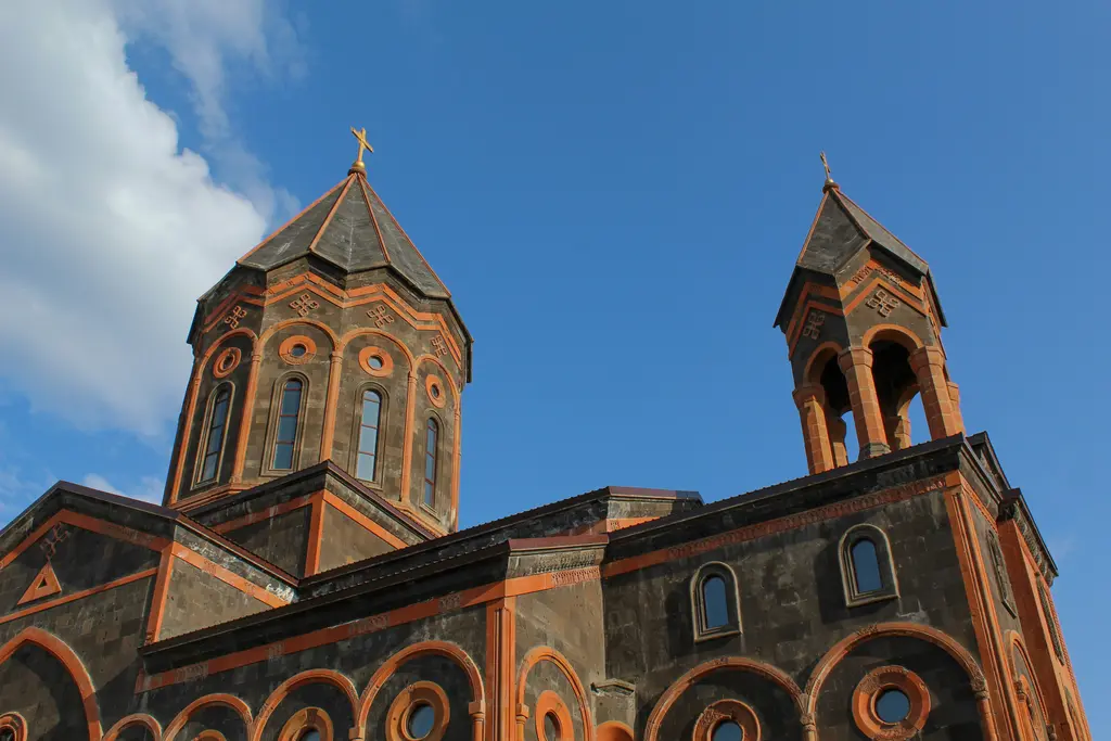 Gyumri'de siyah tüf taşından yapılmış kilisenin üst bölümü; turuncu taşla vurgulanmış kemerler ve haç motifleri, sivri külahlı kubbe kasnağı ve yanında açık kemerli çan kulesi, arkada berrak mavi gökyüzü.