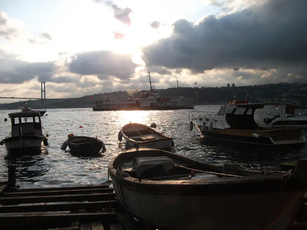Çengelköy kıyısında bağlı kayıklar; bulutlu gökyüzü, parlayan su, vapurlar ve uzakta asma köprü.